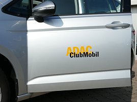 Nahaufnahme einer silbernen Autotür mit der Beschriftung „ADAC ClubMobil“ in gelber und schwarzer Schrift. Im Bild ist der linke Außenspiegel, der Türgriff und ein Teil des Hinterrads zu sehen. Das Fahrzeug steht auf einem gepflasterten Platz. (Foto: Autohaus Peter GmbH )