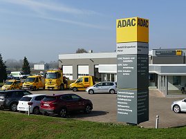 ADAC-Standort in Nordhausen; im Vordergrund eine große gelb-graue ADAC-Stele mit Beschriftung „Mobilitätspartner Peter“. Dahinter mehrere gelbe ADAC-Pannen- und Abschleppwagen und ein modernes, graues Gebäude mit Glasfassade und ADAC-Logo. Im Vordergrund parkende Autos, im Hintergrund Felder und Bäume bei klarem Himmel. (Foto: Autohaus Peter GmbH )