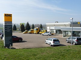 Alt-Text: Blick über das Gelände des ADAC Mobilitätspartners Peter in Nordhausen. Im Vordergrund ein großes gelbes ADAC-Schild mit Serviceleistungen, dahinter mehrere gelbe ADAC-Fahrzeuge und silberne Mietwagen, im Hintergrund das moderne Gebäudekomplex mit Glasfassaden und Werkstatt. (Foto: Autohaus Peter GmbH )