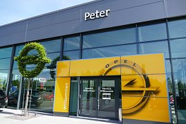 Eingang des Opel-Autohauses Peter in Nordhausen. Über der gläsernen Fassade steht der Schriftzug „Peter“. Vor dem Eingang befindet sich eine große gelbe Wand mit Opel-Logo und „Herzlich Willkommen“-Schriftzug. Links davon steht eine kunstvoll geschnittene grüne Hecke in Form des Opel-Logos. Der Eingangsbereich wirkt modern und einladend. (Foto: Fischer/Autohaus Peter)