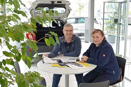 Ein Mann und eine Frau sitzen an einem Tisch im Autohaus, mit Broschüren und Prospekten vor sich, im Hintergrund ein geöffneter Kofferraum eines Autos und helle Glasfronten. (Foto: Fischer/Autohaus Peter)
