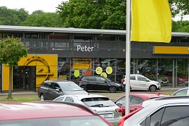 Ansicht des Opel Autohauses Peter mit großen Glasfassaden, gelbem Opel-Logo über dem Eingang und mehreren geparkten Autos im Vordergrund; gelbe Fahne und Bäume im Hintergrund. (Foto: Fischer/Autohaus Peter)