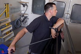 Kurt Kuschmierz beim Entfernen von Dellen an einem Auto, ohne dass lackiert werden muss. Er trägt ein dunkles Polohemd und arbeitet konzentriert mit einem Spezialwerkzeug an der Fahrzeugkarosserie. Im Hintergrund Werkstattausstattung mit Schläuchen, Strahlern und Türen. (Foto: Fischer/Autohaus Peter)