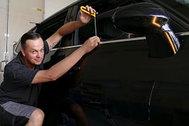 Kurt Kuschmierz arbeitet lächelnd an der Tür eines schwarzen Autos. Er benutzt ein Spezialwerkzeug mit gelbem Griff, um eine Delle vorsichtig auszubeulen. Er kniet neben dem Fahrzeug und zeigt dabei Feingefühl und Präzision. Werkstattumgebung im Hintergrund. (Foto: Fischer/Autohaus Peter)