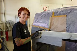 Katja Röpcke führt Schleifarbeiten an einem grundierten Karosserieteil durch. Sie trägt schwarze Handschuhe und ein schwarzes T-Shirt mit Firmenlogo, hat rote Haare und sichtbare Tattoos am Arm. Im Hintergrund ein abgeklebtes Fahrzeug in einer Werkstatt. (Foto: Fischer/Autohaus Peter)