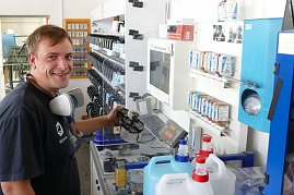 Peter Meyer lächelt, während er an einer Farbmischstation arbeitet. Er hält eine Lackierpistole und trägt eine Atemschutzmaske um den Hals. Vor ihm ein Monitor, viele Farbdosen und große Kanister. Im Hintergrund Regale mit weiteren Farben und Materialien. (Foto: Fischer/Autohaus Peter)