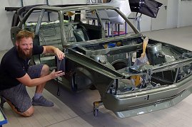 „Tino Schwenzer kniet lächelnd neben einer teilweise montierten grünen Autokarosserie in einer Werkstatt. Er hält ein Messgerät an die Seitenwand, um den Farbton zu bestimmen. Im Hintergrund verschiedene Autoteile und Werkstattausrüstung.  (Foto: Fischer/Autohaus Peter)