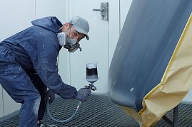 „Marcel Heyer trägt einen blauen Schutzanzug und Atemschutzmaske, während er mit einer Spritzpistole Füller auf ein vorbereitetes Karosserieteil sprüht. Das Teil ist grau grundiert und teilweise mit Papier abgeklebt. Die Szene spielt in einer Lackierkabine. (Foto: Fischer/Autohaus Peter)