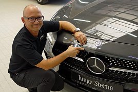 Dirk Morgenthal, Leiter des Lackierzentrums, kniet neben einem schwarzen Mercedes mit dem Schild Junge Sterne. Er trägt ein schwarzes Polohemd mit Aufschrift Peter und und hält ein Farbmessgerät an die Motorhaube, um den exakten Farbton zu bestimmen. Heller Werkstattboden, reflektierende Autolackierung. (Foto: Fischer/Autohaus Peter)