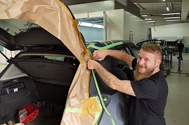 Frank Frank lächelt, während er ein Fahrzeug für Lackierarbeiten mit braunem Abdeckpapier abklebt. Das Auto steht mit geöffneter Heckklappe in einer Werkstatt. Im Hintergrund sind weitere Fahrzeuge und Arbeitsbereiche zu sehen. (Foto: Fischer/Autohaus Peter)