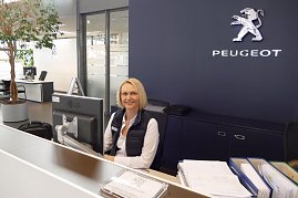 Freundliche Mitarbeiterin am Empfangsschalter eines Peugeot-Autohauses, im Hintergrund das Peugeot-Logo an der Wand, ein PC auf dem Schreibtisch und eine Pflanze links im Bild. (Foto: Fischer/Autohaus Peter)