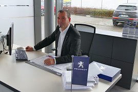 Berater in einem Peugeot-Autohaus sitzt an einem Schreibtisch mit Computer, Unterlagen und einer kleinen blauen Peugeot-Fahne, im Hintergrund Autos vor dem Fenster sichtbar. (Foto: Fischer/Autohaus Peter)