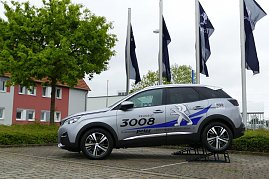 Silberner Peugeot 3008 mit Werbebeschriftung auf einer erhöhten Präsentationsrampe vor einem Autohaus. Im Hintergrund wehen mehrere Peugeot-Flaggen und es steht ein Baum sowie ein Gebäude mit roten und grauen Fassadenelementen. (Foto: Fischer/Autohaus Peter)