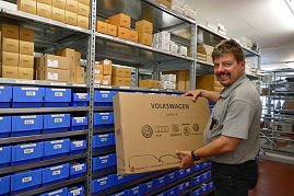 Ein Mitarbeiter in grauem Polohemd steht in einem Ersatzteillager und hält einen großen Karton mit der Aufschrift Volkswagen Group, auf dem Logos von Volkswagen, Audi, Seat, koda und Volkswagen Nutzfahrzeuge abgebildet sind. Im Hintergrund Regale mit blauen Kisten und braunen Kartons. Der Mann schaut lächelnd in die Kamera. (Foto: Fischer/Autohaus Peter)