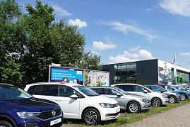 Reihe von VW-Fahrzeugen in unterschiedlichen Farben, die auf einem Parkplatz vor einem modernen Autohaus mit Glasfassade stehen. Im Hintergrund sind Werbetafeln und grüne Bäume zu sehen, der Himmel ist blau mit einigen Wolken. (Foto: Fischer/Autohaus Peter)