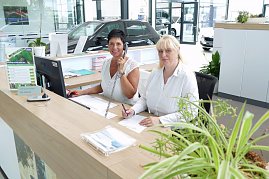 Zwei Serviceassistentinnen sitzen am Empfangstresen in einem modernen Autohaus. Eine Frau telefoniert und lächelt, die andere schreibt etwas auf. Beide tragen weiße Blusen. Auf dem Schreibtisch stehen ein Monitor, Unterlagen sowie Pflanzen. Im Hintergrund sind Autos und der helle Ausstellungsraum zu sehen. (Foto: Fischer/Autohaus Nordhausen)