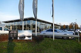 Mercedes-Benz Autohaus mit großer Glasfassade und davor geparkten Fahrzeugen, vor allem silberfarbene Limousinen. Im Vordergrund ein Schild mit der Aufschrift Junge Sterne glänzen länger und einem Bild von Mercedes-Fahrzeugen. Drei hohe Fahnenmasten mit Mercedes-Benz-Fahnen. Grünfläche mit Gras und Sträuchern, blauer Himmel. (Foto: Fischer/Autohaus Peter)