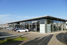 Autohaus Peter mit moderner Glasfassade und der Aufschrift Gebrauchtwagen. Vor dem Gebäude sind mehrere Mercedes-Fahrzeuge in verschiedenen Farben geparkt, darunter ein weißer Wagen im Vordergrund. Weitläufiger Parkplatz, blauer Himmel ohne Wolken. (Foto: Fischer/Autohaus Peter)