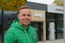 Ihr Verkaufsberater für die amerikanischen Fabrikate: Danny Kindervater mit grüner Jacke steht lächelnd vor dem Jeep-Autohaus, im Hintergrund das Eingangsportal mit Jeep-Logo. (Foto: Fischer/Autohaus Peter)