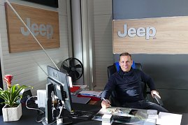 Verkaufsberater Danny Kindervater sitzt an einem Schreibtisch in einem modernen Jeep-Autohaus, mit Jeep-Logos an der Wand, Computer und Pflanzen auf dem Tisch. (Foto: Fischer/Autohaus Peter)
