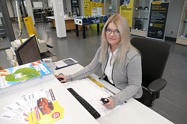 Junge Frau mit langen, blonden Haaren und Brille sitzt am Empfangsschalter des Autohauses. Sie trägt einen grauen Cardigan über einer weißen Bluse und lächelt leicht in die Kamera. Auf dem Schreibtisch liegen Prospekte, ein Monitor steht vor ihr. Im Hintergrund sind Verkaufs- und Beratungsbereiche des Opel-Autohauses sichtbar. (Foto: Fischer/Autohaus Peter)