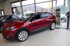 Roter Opel Grandland X Hybrid im Innenraum des Opel-Autohauses, während er an einer Ladestation angeschlossen ist. Das Fahrzeug steht vor großen Glasfenstern, durch die man Häuser und Hügel im Hintergrund sieht. Auf der Fahrerseite ist die Aufschrift „Grandland X Hybrid“ sichtbar. Daneben weitere Fahrzeuge und ein hell gefliester Boden. (Foto: Fischer/Autohaus Peter)