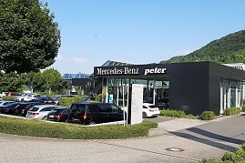 Außenansicht des Mercedes-Benz Autohauses Peter in Sondershausen. Das moderne Gebäude mit schwarzer Fassade trägt die Aufschrift 'Mercedes-Benz' und 'Peter'. Davor sind viele Autos auf dem Parkplatz geparkt, unter anderem ein schwarzer Van. Im Hintergrund sieht man grüne Bäume und einen bewaldeten Hügel unter blauem Himmel. (Foto: Fischer/Autohaus Peter)