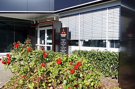 Eingangsbereich eines Mitsubishi-Autohauses mit moderner Glas- und Metallfassade. Davor blühende rote Rosen und ein schwarzes Schild mit Öffnungszeiten und rotem Mitsubishi-Logo. (Foto: Fischer/Autohaus Peter)