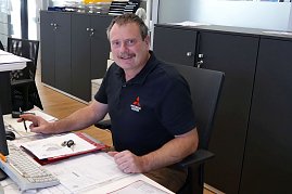 Freundlicher Mitarbeiter in Mitsubishi-Motors-Polo sitzt an einem Schreibtisch im Büro, hält einen Stift in der Hand und lächelt in die Kamera. (Foto: Fischer/Autohaus Peter)