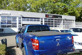 Blauer Mitsubishi-Pick-up vor der Mitsubishi-Servicewerkstatt mit großen Glasgaragentoren und Mitsubishi-Motors-Logo, Bäume im Hintergrund. (Foto: Fischer/Autohaus Peter)