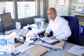 Mann in weißem Hemd sitzt an einem Schreibtisch im Büro, schaut in die Kamera, schreibt in einen Terminplaner. Auf dem Tisch liegen viele Unterlagen, ein Computer, Tastatur, Kaffeetasse und Büromaterial. (Foto: Fischer/Autohaus Peter)