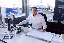 Autohaus-Mitarbeiter in weißem Hemd sitzt an einem Schreibtisch vor einem Computer, lächelt in die Kamera, Verkaufsunterlagen und Broschüren auf dem Tisch, Autos sind draußen durch das Fenster sichtbar. (Foto: Fischer/Autohaus Peter)