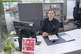 Mitarbeiter im Autohaus sitzt lächelnd an einem Schreibtisch mit Computer, Unterlagen und Pflanze, im Hintergrund Regale und Fenster mit Blick nach draußen. (Foto: Fischer/Autohaus Peter)