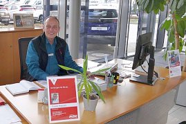 Ein Mitarbeiter sitzt freundlich lächelnd an einem Schreibtisch im Autohaus Peter in Göttingen, mit Namensschild und Pflanzen auf dem Tisch, im Hintergrund ein Auto mit Peter-Nummernschild. (Foto: Fischer/Autohaus Peter)