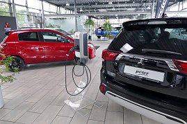 „Innenraum eines Autohauses mit mehreren Mitsubishi-Fahrzeugen, im Vordergrund eine Ladestation für Elektrofahrzeuge zwischen einem roten Kleinwagen und einem schwarzen SUV. (Foto: Fischer/Autohaus Peter)