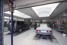 Helle Mercedes-Benz Werkstatt mit zwei Fahrzeugen auf Hebebühnen: links ein schwarzer Geländewagen (G-Klasse), rechts ein silberner Oldtimer. Im Hintergrund Glasbüros und Regale mit Autopflegeprodukten. (Foto: Fischer/Autohaus Peter)