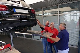 Drei Männer stehen in einem hellen Werkstattbereich unter dem Heck eines schwarzen Mercedes-AMG, der auf einer Hebebühne angehoben ist. Einer der Männer zeigt auf eine Stelle am Fahrzeug, die anderen beiden schauen interessiert zu. (Foto: Fischer/Autohaus Peter)