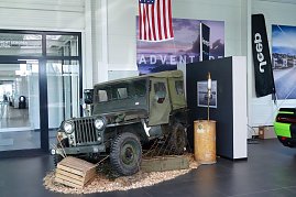 Historischer Jeep Willy in nostalgischer Präsentation im Showroom mit US-Flagge und Jeep-Banner im Hintergrund. (Foto: Fischer/Autohaus Peter)