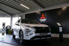 Weißer Mitsubishi Eclipse Cross Plug-in Hybrid glänzend in einem Showroom ausgestellt, mit großem schwarzen Hintergrund und rotem Mitsubishi Motors Logo an der Wand. (Foto: Fischer/Autohaus Peter)