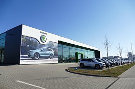 koda-Autohaus mit großer Glasfassade und grünem Eingang, daneben viele geparkte Autos in einer Reihe, vorne ein großes Werbeplakat mit einem koda-SUV, blauer Himmel. (Foto: Fischer/Autohaus Peter)