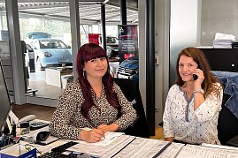 Zwei Mitarbeiterinnen sitzen an einem Empfangstresen im Autohaus, eine telefoniert freundlich, die andere füllt Unterlagen aus. Im Hintergrund stehen Autos in einem hellen Ausstellungsraum. (Foto: Fischer/Autohaus Peter)