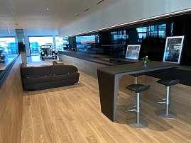 Moderner Kundenwartebereich im Autohaus mit Holzfußboden, dunkelbraunem Sofa, Hochtisch mit Barhockern, historischen Fotos und Blick auf ein klassisches Auto im Hintergrund. (Foto: Arenswald / Autohaus Peter)