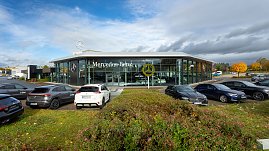 Mercedes-Benz Autohaus mit moderner Glasfassade und großem Mercedes-Logo, mehrere geparkte Fahrzeuge im Vordergrund, herbstliche Bäume und Wolkenhimmel im Hintergrund. (Foto: Steve Dornhofer/Autohaus Peter)
