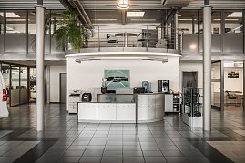 Moderner Empfangsbereich eines Autohauses mit halbkreisförmigem Tresen, Druckern, Kaffeemaschine und Wasserspender. An der Wand hängt ein Bild mit Mercedes-Autos. Darüber eine Galerie mit Geländer und Tisch mit Stühlen. Der Raum hat große Fenster, graue Fliesen und eine hohe Decke. (Foto: Eric Freytag)