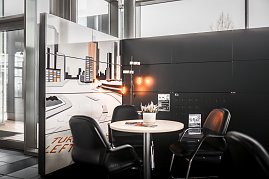 Gemütliche Sitzecke mit rundem Tisch und vier schwarzen Lederstühlen in einem Autohaus. Auf dem Tisch steht eine kleine Pflanze. Im Hintergrund eine Wand mit großem Stadtmotiv in Weiß- und Orangetönen. Helle Fensterfront und moderner schwarzer Wandbereich. (Foto: Eric Freytag)