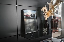 „Dekoratives Regal mit einem gerahmten Junge Sterne-Zertifikat von Mercedes-Benz, daneben zwei Vasen mit Trockenblumen in Braun- und Beigetönen. Auf dem Regal liegen auch Magazine, davor eine Glaskugel-Deko. Moderner schwarzer Hintergrund. (Foto: Eric Freytag)