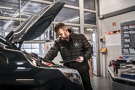 „Werkstattmitarbeiter im Mercedes-Benz Autohaus prüft den Motorraum eines Fahrzeugs mit geöffnetem Vorderhaube. Er trägt eine schwarze Jacke mit Mercedes-Logo und hält ein Tablet. Im Hintergrund moderne Werkstatteinrichtung mit Dokumentenfächern, Kalender und Glastüren. (Foto: Eric Freytag)