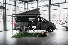 „Grauer Mercedes-Benz Camper-Van mit ausgefahrener Markise und aufgeklapptem Schlafdach in einer hellen Ausstellungshalle. Vor dem Van stehen zwei Campingstühle und ein Tisch auf grünem Teppich, daneben ein dekorativer Korb mit Holzscheiten. Draußen ist Schnee zu sehen. (Foto: Eric Freytag)
