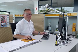 Ronny Peukert, Verkaufsberater, sitzt freundlich lächelnd an einem modernen Schreibtisch im hellen Autohaus-Büro. Er trägt ein weißes Hemd und eine Brille. Auf dem Schreibtisch stehen ein Monitor, Tastatur, Maus, Schreibtischlampe und dekorative Elemente mit Opel-Logo. Im Hintergrund sind Schränke und Büromaterialien zu sehen. (Foto: Autohaus Peter Gruppe)