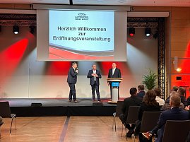 Nahaufnahme der Bühne: Drei Männer im Anzug (rechts Helmut Peter) stehen vor einer Leinwand mit der Begrüßung zur Eröffnungsveranstaltung. Einer spricht ins Mikrofon. (Foto: Autohaus Peter )